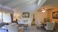 This single-floor residence makes a striking impression with its dramatic vaulted ceiling, finished in white-painted tongue-and-groove wood planking that rises to a generous peak and is punctuated by a high clerestory window that draws natural light deep into the interior. Exposed beams and a ceiling fan add both character and practicality to the airy space.

The open-concept layout seamlessly connects the living and dining areas, creating a cohesive and inviting atmosphere. The living area is anchored by a plush white sofa and coordinating armchairs arranged around a glass-topped coffee table with ornate ironwork legs. A decorative gilt-framed mirror adorns the wall, complemented by warm ambient lighting from classic table lamps and an elegant tall copper vase with dried botanicals that adds a sculptural accent.

The dining area transitions naturally from the living space and features a round table surrounded by tufted, upholstered chairs, lending a refined and classic sensibility to the room. Neutral carpet throughout ties the spaces together while maintaining a soft, warm tone. The overall ambiance is bright, sophisticated, and welcoming, with a careful blend of traditional furnishings and architectural character that gives the home a timeless, comfortable elegance.