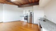 Located in the heart of Richmond, Virginia, this single-floor loft-style unit at 12 S 17th St showcases a compelling blend of industrial heritage and contemporary updates. The space is defined by its striking exposed brick walls, which run throughout and lend an authentic, historic character to the interior. Overhead, rich wooden plank ceilings with prominent structural beams add warmth and architectural depth, creating a distinctive loft aesthetic.

The kitchen is well-appointed with white shaker-style cabinetry, granite countertops, and a full suite of stainless steel appliances, including a refrigerator, electric range, and dishwasher. Warm hardwood flooring flows through the main living and kitchen areas, complementing the natural tones of the wood ceiling.

The bedroom features plush carpeting and continues the exposed brick motif, with a closet and a window that brings in natural light. Functional black piping is visible along the brick walls, reinforcing the industrial character of the space. The bathroom is clean and utilitarian, equipped with a stand-up shower and toilet.

Natural light filters in through multiple windows along the brick walls, contributing to an open, airy feel throughout. This space offers a unique opportunity to experience Richmond's architectural history paired with modern-day comforts.