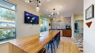This single-story amenity space is thoughtfully designed to serve as a community hub, offering distinct zones for socializing, working, and exercising. The co-working and lounge area is anchored by a substantial butcher-block communal table surrounded by industrial-style metal bar stools, set beneath a striking multi-globe pendant chandelier. Large windows along the perimeter flood the space with natural light and offer views of the surrounding greenery. Light hardwood flooring runs throughout, complementing the warm, neutral-toned walls accented by exposed brick detailing near a beverage station outfitted with a coffee maker and counter space. A wall-mounted flat-screen television adds a functional, modern touch.

The adjacent lounge features a teal sofa, modern accent chairs, patterned ottomans, and a large inspirational quote mural by George Bernard Shaw, lending the area an intellectual and welcoming character. Recessed lighting and a contemporary chandelier complete the ambiance.

The fitness center is divided into two well-appointed sections: a cardio zone lined with treadmills and ellipticals alongside floor-to-ceiling mirrors, and a strength training area featuring cable machines, free weights, and resistance equipment, all set against a rustic wood-paneled accent wall branded with a "Trade Up" logo.