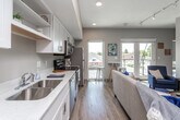 This single-floor unit offers a thoughtfully designed open-concept layout that seamlessly connects the kitchen and living area, creating a bright and functional living space. Large windows and a glass door flood the interior with natural light while offering views of the surrounding neighborhood, enhancing the airy ambiance throughout.

The kitchen is outfitted with crisp white shaker-style cabinetry, light quartz countertops, and a full suite of stainless steel appliances, including a refrigerator, electric range, over-the-range microwave, and dishwasher. Open shelving adds a modern, accessible touch to the cabinetry arrangement. The living area features elevated ceilings with track lighting, providing a sense of openness, and includes a dedicated wall mount for a television.

A tall bistro-style bar table positioned near the large windows offers a casual dining option with an outdoor view. The bedroom is a calm, neutral-toned retreat with warm hardwood-style flooring and ample wall space. The bathroom is well-appointed with a dark-finished vanity, light countertop, a tub-shower combination, and hardwood-style flooring that ties the space together cohesively. Throughout the unit, neutral wall tones and consistent light flooring create a clean, modern aesthetic.