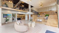 This expansive, thoughtfully designed lobby makes an immediate impression with its soaring double-height ceilings and dramatic floor-to-ceiling glass curtain walls that flood the interior with natural light and offer sweeping views of the surrounding streetscape. The architectural design masterfully contrasts warm natural materials — including light oak wood flooring, wood-paneled built-in seating, and richly textured accent walls — against raw exposed concrete columns and dark, industrial-style ceiling elements.

The main lounge area anchors the space with a curved, blush-toned sectional sofa, a patterned area rug, and organic wood stump accent tables, creating an inviting and stylish focal point. Statement sputnik chandeliers in brushed gold add a bold artistic touch overhead. A sleek, curved reception desk with a matte dark finish sits within a secondary area featuring an exposed black ceiling with globe and pendant lighting fixtures, white cabinetry, and glass-partitioned private rooms beyond.

A striking open staircase with light wood treads and black metal railings connects to an upper mezzanine level, where additional lounge and common areas are visible. Built-in bench seating lines a hallway adorned with a textured, macramé-style wall installation. The space flows seamlessly across multiple connected hallways and zones, balancing a warm, welcoming ambiance with a polished, contemporary aesthetic throughout.