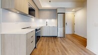 Located in Toronto, Ontario, this modern single-floor unit offers four bedrooms and one bathroom within a thoughtfully designed open layout. The kitchen and living area flow seamlessly together, anchored by light wood-look flooring that extends throughout the entire unit. The contemporary kitchen features light wood-grain cabinetry extending to the ceiling, a clean white countertop and backsplash, stainless steel appliances including a built-in microwave and range, an integrated refrigerator, and a undermount sink with a matte black faucet. Track lighting accentuates the kitchen space. The living area is bright and airy, benefiting from large floor-to-ceiling windows that frame views of the surrounding Toronto skyline, flooding the space with natural light. The bedrooms are well-proportioned, with the generously sized primary bedroom offering ample space. Several bedrooms also feature expansive glazed windows with city views. A hallway with mirrored sliding closet doors connects the private areas of the unit. The bathroom is finished with large-format marble-effect grey tiles covering the walls and floor, complemented by a walk-in shower with a white tray base, a floating vanity, and matte black hardware, creating a refined and spa-like atmosphere.
