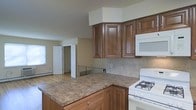 This single-floor space offers a practical and inviting layout centered around a well-appointed kitchen. The kitchen is a standout feature, showcasing rich, warm-toned wood cabinetry with raised panel doors that extend to the upper walls, providing generous storage. A central island with a granite-style countertop serves as both a workspace and a gathering point, adding both function and visual appeal. White appliances, including a gas range and an over-the-range microwave, complement the neutral aesthetic. A classic tile backsplash ties the cooking area together with a clean, timeless look. The open layout allows the kitchen to flow naturally into the adjoining living area, which benefits from natural light streaming through a window. Light hardwood-style flooring runs throughout, contributing to a warm and cohesive feel. A staircase railing visible from the kitchen hints at additional access points, and interior doors lead to separate rooms, offering defined spaces within the overall open feel. Neutral wall tones throughout create a versatile backdrop suitable for a variety of furnishing styles. The space is designed for comfortable, everyday living with an emphasis on practicality and ease of movement between areas.