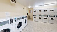 This shared laundry facility offers a practical and efficiently organized space designed to accommodate high-volume laundry needs. The room is equipped with a row of front-loading washing machines along one wall, providing ample capacity for simultaneous use. Opposite the washers, multiple stacked dryer units are arranged to maximize the available floor space, offering a generous number of drying cycles at once. A dedicated folding table is conveniently positioned near the machines, providing a sturdy surface for sorting and folding laundry. The flooring throughout is durable terrazzo, featuring a speckled white pattern that is both resilient and easy to maintain. The walls are finished in a clean, neutral cream tone, contributing to a bright and hygienic atmosphere. Overhead fluorescent lighting ensures the room is well-lit at all times. A row of clerestory windows along the upper portion of one wall allows natural light to supplement the artificial lighting. Informational notices and posted signage are displayed near the entry area, and a wall-mounted clock provides easy time reference. The room also includes baseboard heating and a ventilation unit, ensuring comfortable conditions year-round. The overall layout is open and easy to navigate, making this laundry room a functional and user-friendly amenity.