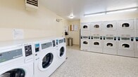 This shared laundry facility offers a practical and efficiently organized space designed to accommodate high-volume laundry needs. The room is equipped with a row of front-loading washing machines along one wall, providing ample capacity for simultaneous use. Opposite the washers, multiple stacked dryer units are arranged to maximize the available floor space, offering a generous number of drying cycles at once. A dedicated folding table is conveniently positioned near the machines, providing a sturdy surface for sorting and folding laundry. The flooring throughout is durable terrazzo, featuring a speckled white pattern that is both resilient and easy to maintain. The walls are finished in a clean, neutral cream tone, contributing to a bright and hygienic atmosphere. Overhead fluorescent lighting ensures the room is well-lit at all times. A row of clerestory windows along the upper portion of one wall allows natural light to supplement the artificial lighting. Informational notices and posted signage are displayed near the entry area, and a wall-mounted clock provides easy time reference. The room also includes baseboard heating and a ventilation unit, ensuring comfortable conditions year-round. The overall layout is open and easy to navigate, making this laundry room a functional and user-friendly amenity.
