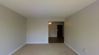 This single-floor space presents a straightforward, functional layout characterized by clean lines and neutral finishes throughout. The main living area is finished with wall-to-wall carpeting in a warm beige tone, complemented by smooth white ceilings and light gray-beige walls that create a calm, understated atmosphere. White baseboards provide a crisp border along the perimeter of the room.

An open passageway connects the main living area to an adjoining room, which features contrasting dark hardwood-style flooring. This secondary space is equipped with a ceiling fan, offering both lighting and air circulation. A dark entry door is visible in the background, suggesting this connected area may serve as a dining space or entryway leading further into the unit.

The walls are relatively bare, with light switch plates and electrical outlets positioned at standard heights, indicating a move-in-ready condition. The overall design is modest and practical, with the open sightlines between rooms contributing to a sense of connected, flowing space. The neutral palette throughout provides a versatile backdrop suitable for a variety of furnishing styles.