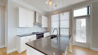 Located at 1414 Broadview Ave in Columbus, Ohio, this single-floor home offers a thoughtfully designed interior with modern finishes throughout. The kitchen is a standout feature, showcasing white shaker-style cabinetry that extends to the ceiling, complemented by a striking marble-style subway tile backsplash. Dark quartz countertops provide a bold contrast against the bright cabinetry, adding a contemporary edge to the space. A central island with an undermount sink serves as a functional focal point, ideal for meal preparation and casual gatherings. Stainless steel appliances, including a freestanding range with a sleek wall-mounted hood, complete the culinary setup. Pendant lighting with warm-toned Edison bulbs hangs above the island, adding an inviting ambiance. Large windows dressed with horizontal blinds allow generous natural light to fill the kitchen, creating a bright and airy atmosphere. Light hardwood flooring runs throughout, tying the space together with warmth and continuity. The layout is efficient and open, offering a smooth flow between areas. Crown molding adds a refined architectural detail, elevating the overall design of this well-appointed Columbus home.