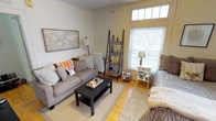 This single-floor space offers a charming and functional living environment defined by its classic architectural character. The living room serves as the heart of the space, showcasing crown molding, hardwood floors, and a large double-hung window with a transom above that floods the room with natural light and frames a pleasant outdoor view of mature trees. A traditional white cast-iron radiator adds a period-appropriate detail that complements the room's timeless aesthetic. The space is thoughtfully arranged with comfortable seating, a central coffee table, and a lean ladder-style bookshelf that provides both storage and display space for books and decorative items. A framed world map anchors one wall, adding a sense of personality and depth to the room. Soft, neutral tones throughout — from the light gray upholstery to the layered area rug — create a warm and inviting ambiance. The overall design strikes a balance between casual comfort and curated style, making the living area feel both livable and welcoming. The abundance of natural light and the classic architectural details give the space a distinctive character well-suited for everyday living.