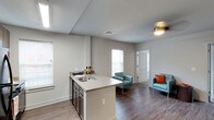 This single-floor unit showcases an open-concept design that seamlessly connects the kitchen and living areas. The kitchen features a peninsula with a built-in sink and dark cabinetry below, providing both functional workspace and a natural divider between the two zones. Light-toned countertops contrast with the dark wood-style cabinetry, creating a clean, modern aesthetic.

The flooring throughout is a rich, dark hardwood-style material that adds warmth and continuity across the space. Neutral light gray walls keep the interior feeling fresh and versatile. Multiple windows fitted with white horizontal blinds allow generous natural light to fill the room, enhancing the open and airy atmosphere.

The living area is well-proportioned, accommodating seating comfortably, as demonstrated by the teal accent chairs with colorful throw pillows visible in the space. A ceiling fan with a brushed gold fixture provides both lighting and air circulation. A white paneled door leads to an adjacent room or exterior access point, adding to the functional flow of the layout. Overall, the unit offers a clean, contemporary feel with practical finishes throughout.