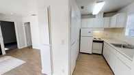 This single-floor space offers a straightforward and functional layout designed for practical everyday living. The kitchen is equipped with essential white appliances, including a full-size refrigerator and an electric range, complemented by white cabinetry that provides ample storage. Overhead fluorescent lighting keeps the kitchen area well-lit, while a ventilation duct ensures proper airflow. The wood-look laminate flooring runs throughout the space, adding warmth and continuity to the interior. The walls are finished in clean white throughout, lending a bright and open feel to the overall environment. A partial wall and doorway arrangement creates a natural division between the kitchen and the adjoining living or hallway area, offering a sense of separation without sacrificing openness. The space maintains a neutral, no-frills aesthetic that provides a clean canvas for personalization. Accessible electrical outlets and light switches are conveniently positioned along the walls. Overall, the space presents a compact yet efficient floor plan suited for comfortable, everyday use.