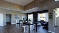 This single-floor space showcases a contemporary design with a clean, modern aesthetic. The room features notably high ceilings accented by recessed lighting, creating an open and airy atmosphere. The two-tone wall treatment — warm gray upper walls transitioning to a bold dark accent wall — adds visual depth and sophistication throughout the space.

The layout is thoughtfully divided into distinct functional zones. A central gathering area is anchored by sleek black bar-height tables paired with modern white swivel stools, ideal for casual meetings or collaborative work. Adjacent to this, a comfortable lounge area features a charcoal upholstered chair and ottoman arranged around a low circular side table, providing a more relaxed setting.

Large windows dressed with white curtains line one wall, allowing generous natural light to flood the interior while offering views of the surrounding greenery. Dark hardwood-style flooring grounds the space and complements the overall color palette. Additional details include wall-mounted artwork, a flat-screen TV on the accent wall, and subtle decorative greenery that adds warmth. A workstation area along one wall provides a functional touch, making this a versatile, well-appointed communal space.