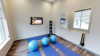 This single-floor fitness and wellness studio is thoughtfully designed to support a range of mind-body exercise practices. The space features neutral gray walls and wood-look flooring, creating a clean and calming environment conducive to focused movement and relaxation. Blue yoga mats are laid out across the floor, accompanied by stability balls ready for use in core and balance training. A vertical rack holds a variety of medicine balls, providing options for strength and functional fitness exercises. A classic ballet barre runs along one wall beneath a large mirror, ideal for barre workouts, stretching, and alignment practice. The mirror enhances the sense of openness within the room while allowing practitioners to monitor their form. A wall-mounted flat-screen display is positioned for easy viewing during guided workout sessions or instructional content. A window on the adjacent wall allows natural light to filter into the space, adding to the bright and airy atmosphere. The studio's layout is open and uncluttered, maximizing usable floor space for group or individual sessions. Overall, the space is well-equipped and purposefully arranged to support yoga, Pilates, barre, and general fitness activities in a welcoming, professional setting.