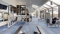 Located at 6880 W 91st Ct in Westminster, Colorado, this well-appointed fitness center offers a comprehensive workout environment designed with both functionality and aesthetics in mind. The facility is housed within a striking architectural space featuring soaring ceilings, angled glass walls, and a greenhouse-style glazed roof section that floods the interior with natural light and provides open views of the surrounding Colorado landscape.

Upon entry, a tiered staircase with sleek metal railings leads down into the main workout floor, finished with durable blue-toned carpet tile throughout. The cardio zone is lined with a row of treadmills and elliptical machines positioned along the expansive glass wall, allowing users to exercise with panoramic outdoor views. Ceiling fans circulate air throughout the space, enhancing comfort during workouts.

The strength training area is generously equipped with a full rack of dumbbells, medicine balls, flat and adjustable benches, cable machines, and multi-use weight equipment. Wall-mounted mirrors amplify the sense of space and support proper form during exercise. A mounted flat-screen television is also present for added convenience. The open layout provides ample room to move between stations, making this fitness center a well-rounded and inviting space for all training styles.