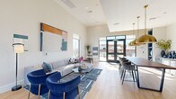 Located at 9944 Highway 6 in Missouri City, TX, this single-floor leasing and welcome center for Inspire Homes offers a thoughtfully designed, open-concept space that combines a living area and dining zone into one cohesive environment. The interior is immediately distinguished by its soaring ceilings and expansive floor-to-ceiling windows and glass doors, which flood the space with natural light and provide clear views of the outdoor pool and community grounds beyond.

Light wood-look flooring runs throughout, complementing the neutral, warm white walls and creating a bright, welcoming atmosphere. The living area features a contemporary lounge setup with a channeled gray sofa, bold navy accent chairs with gold-toned legs, and a patterned area rug — all anchored by a large abstract artwork on the wall. A decorative frosted glass partition wall with an intricate geometric pattern adds an elegant architectural detail near the entry.

The dining zone is defined by a warm wood table beneath a striking brass dome pendant light, with a built-in blue display shelf unit and a marble-style backsplash counter area adding character and depth. Glass-paneled interior doors with black frames lead to private leasing offices, maintaining visual openness while delineating functional areas. The entry features branded Inspire Homes signage and access to the outdoor community amenities.