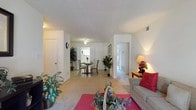 This single-floor residence offers a comfortable and functional layout designed for easy living. The open-plan living area serves as the heart of the home, featuring light-toned carpeting, white walls, and a textured ceiling that adds subtle visual interest. A vibrant red area rug anchors the living space, which is furnished with a classic sofa and accented by a warm table lamp and decorative floral arrangement, creating an inviting atmosphere. Lush indoor plants are thoughtfully placed throughout, adding a refreshing, natural element to the interior. The living area flows seamlessly into the dining space, which features a round dining table with chairs positioned beneath a ceiling light fixture. The tile flooring in the dining area offers a clean transition from the carpeted living zone. Beyond the dining area, a glimpse into the kitchen reveals bright, well-lit cabinetry and workspace. A hallway visible from the main living area leads to additional rooms, with natural light streaming through, suggesting well-placed windows throughout. The overall ambiance is light, airy, and welcoming, with a neutral color palette that provides a versatile backdrop for personalization.
