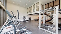 Located at 5600 E 84th Ter in Kansas City, MO, this fitness center offers a thoughtfully designed exercise environment across a single, generously proportioned floor. The space is defined by its dramatic double-height ceilings and expansive floor-to-ceiling windows, which flood the main workout area with abundant natural light while offering lush outdoor views of surrounding greenery and a glimpse of a pool area beyond.

The main exercise floor features durable rubberized flooring throughout and is outfitted with a comprehensive selection of cardio equipment, including multiple treadmills and elliptical trainers, as well as a multi-station strength machine and a rack of dumbbells and medicine balls for free-weight training. A striking open-riser staircase with wood treads and steel railings adds an architectural focal point to the space, leading to an upper-level mezzanine area that overlooks the main floor.

A dedicated functional training room provides space for high-intensity workouts, equipped with a battle rope station, a large training tire, and additional conditioning equipment set against clean white walls and recessed lighting. The overall ambiance is modern and motivating, combining sleek design elements with a practical, full-featured fitness facility suitable for a wide range of training styles.