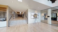 This single-floor lobby presents a refined, modern aesthetic anchored by large-format polished stone tile flooring that extends throughout the entire space. The entrance is defined by a set of bold black-framed glass doors flanked by expansive floor-to-ceiling windows, flooding the interior with abundant natural light and offering clear sightlines to the surrounding exterior. Upon entry, the space opens dramatically, showcasing soaring ceilings that amplify the sense of grandeur and volume.

A sculpted white reception desk is prominently positioned near the core of the lobby, set against a warm wood-paneled backdrop with integrated lighting that adds depth and texture. Stone-clad accent walls complement the neutral palette throughout. A raised corridor, accessed by a broad tiled staircase with a sleek metal railing, connects the main entry area to a deeper hallway, lending the layout a dynamic sense of layered elevation.

The lounge area is generously proportioned, featuring multiple seating arrangements with teal and charcoal patterned armchairs, deep sofas, and occasional tables arranged atop colorful area rugs. Decorative artwork and accent lighting enhance the polished, welcoming atmosphere, making the space well-suited for both arrivals and social interaction.
