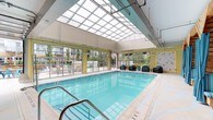 This amenity space centers around a well-appointed indoor swimming pool bathed in natural light streaming through an expansive overhead skylight that spans the length of the pool enclosure. Floor-to-ceiling glass walls and large garage-style doors open directly to an outdoor courtyard, seamlessly blending the indoor and outdoor environments. The pool area features light yellow tile walls, a painted concrete deck, and rattan lounge chairs with teal cushions positioned poolside for relaxed seating. Adjacent to the main pool, a blue mosaic-tiled hot tub spa occupies its own alcove, accompanied by a wooden privacy partition with turquoise curtains, pendant lighting, and decorative wall art, creating a more intimate atmosphere. Water fountains and standard safety amenities are also present within the pool facility.

The outdoor courtyard beyond is a generously scaled gathering space with a bold geometric tile pattern on the patio floor. A central circular seating area with a fire pit is surrounded by rocking chairs and side tables, while nearby communal dining tables with colorful metal chairs offer additional gathering options. String lights overhead, a bar area, and lush plantings throughout add character and vibrancy to the space.