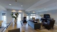 This thoughtfully designed single-floor space showcases a seamless open-concept layout that effortlessly connects the dining and living areas, creating a generous and welcoming atmosphere. The interior is anchored by a sleek, modern double-sided fireplace that serves as a stunning focal point, elegantly dividing the space while maintaining visual continuity throughout.

The living area is furnished with a plush grey sofa, complementary accent chairs, and a flat-screen television, all arranged to encourage comfort and conversation. Recessed lighting throughout the ceiling provides warm, layered illumination that enhances the space's sophisticated ambiance. Large windows dressed with dark Roman shades allow abundant natural light to flood the interior while offering a view of the outdoors.

The dining area features a long wooden table paired with sleek seating, accented by tall decorative planters with lush greenery that bring a touch of nature indoors. The light-toned hardwood flooring laid in a classic herringbone pattern adds a refined, timeless elegance underfoot and ties the entire space together cohesively. The neutral color palette of whites, greys, and warm wood tones creates a calm, contemporary aesthetic that feels both polished and inviting throughout.