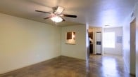 This single-story space features an open-concept layout that connects the living area to the kitchen, creating a functional and cohesive floor plan. The living area is finished with polished concrete floors, which lend an industrial yet practical character to the space. A ceiling fan with an integrated light fixture is centrally mounted, providing both airflow and ambient lighting overhead.

A distinctive architectural detail is the pass-through window cut into the wall between the living area and the kitchen, fitted with a countertop ledge and accented by a pendant light. This design element enhances connectivity between the two spaces while maintaining a degree of separation.

The kitchen is equipped with dark cabinetry, a stainless steel refrigerator, and what appears to be a stacked washer and dryer unit conveniently integrated into the layout. A window in the kitchen area allows natural light to filter in. The walls throughout are painted in a neutral off-white tone, contributing to a clean and understated aesthetic. Overall, the space offers a straightforward, utilitarian design with practical features suited for everyday living.