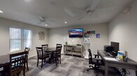 This single-floor communal space is thoughtfully designed to serve as a shared gathering and work area. The main room is finished with neutral gray walls and patterned carpet tiles in varying shades of gray, lending a clean, modern aesthetic throughout. Two ceiling fans with integrated lighting provide comfortable air circulation and ambient illumination, complemented by recessed lighting in the ceiling.

The seating area is furnished with two round dark wood dining tables, each surrounded by matching dark wood chairs, offering ample space for group gatherings or casual meetings. A wall-mounted flat-screen television is positioned above a mirrored console table, creating a focal point for the lounge area. Decorative metallic wall art adds a contemporary touch to the otherwise understated décor.

Adjacent to the lounge, a dedicated computer workstation features a dark wood desk, an ergonomic office chair, a desktop computer, and a printer — providing residents or guests with a practical workspace. Large windows dressed with vertical blinds allow natural light to filter into the room.

A doorway connects the main space to a kitchenette, which is visible in the background and equipped with cabinetry and essential appliances, adding convenience to the overall layout. The space strikes a balance between comfort and functionality.
