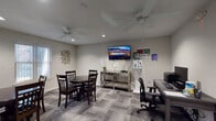 This single-floor communal space is thoughtfully designed to serve as a shared gathering and work area. The main room is finished with neutral gray walls and patterned carpet tiles in varying shades of gray, lending a clean, modern aesthetic throughout. Two ceiling fans with integrated lighting provide comfortable air circulation and ambient illumination, complemented by recessed lighting in the ceiling.

The seating area is furnished with two round dark wood dining tables, each surrounded by matching dark wood chairs, offering ample space for group gatherings or casual meetings. A wall-mounted flat-screen television is positioned above a mirrored console table, creating a focal point for the lounge area. Decorative metallic wall art adds a contemporary touch to the otherwise understated décor.

Adjacent to the lounge, a dedicated computer workstation features a dark wood desk, an ergonomic office chair, a desktop computer, and a printer — providing residents or guests with a practical workspace. Large windows dressed with vertical blinds allow natural light to filter into the room.

A doorway connects the main space to a kitchenette, which is visible in the background and equipped with cabinetry and essential appliances, adding convenience to the overall layout. The space strikes a balance between comfort and functionality.
