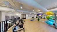 Located at 2395 Herodian Way SE in Atlanta, GA, this contemporary fitness studio offers a well-appointed, open-plan workout environment designed to support a wide range of exercise disciplines. The space features light hardwood flooring throughout, contributing to a clean and energizing atmosphere, while exposed white ductwork along the elevated ceilings adds an industrial-modern character.

The studio is generously equipped with a row of indoor cycling bikes, WaterRower rowing machines, and an assortment of stability balls in multiple sizes displayed on vertical racks. A large wall-to-wall mirror along one side visually expands the space and serves as a functional training aid. A wall-mounted display screen supports guided workouts and class programming.

Natural light pours in abundantly through expansive floor-to-ceiling windows along the exterior walls, offering views of the surrounding building complex. Exposed concrete columns punctuate the space, reinforcing the architectural character. Framed artwork on the walls adds a refined, curated touch to the environment.

Access to the studio is through sleek black-framed glass double doors, which lend a sophisticated, boutique-gym aesthetic. The open layout provides ample floor space for mat-based exercises, yoga, and stretching, making this facility versatile and accommodating for a variety of fitness routines.