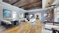 This elegant single-floor space makes an immediate impression with its distinctive coffered ceiling finished in rich, dark wood planking, accented by recessed lighting that casts a warm, inviting glow throughout the open interior. Wide-plank hardwood flooring grounds the space with a natural warmth that complements the refined design palette above.

The main lounge area is generously proportioned, featuring multiple seating arrangements with contemporary white upholstered sofas and accent chairs arranged to encourage both social gatherings and relaxed conversation. A bold, marbled textured feature wall in deep blue-grey tones serves as a striking focal point, anchoring a mounted flat-screen display and built-in bar or beverage area with shelving above.

Large windows along one side of the space flood the interior with natural light, enhancing the bright, airy atmosphere. A counter-height bar seating area provides an additional casual gathering spot, while an adjoining room visible through a wide doorway extends the functional footprint and offers further lounge or meeting space. Decorative wallcoverings and carefully curated mirrors and artwork add layers of sophistication throughout. The overall ambiance is upscale and welcoming, blending modern design elements with warm, residential-inspired finishes.