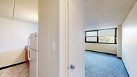 This one-bedroom, one-bathroom unit at 9 Lamont Ave in Trenton, NJ offers a practical and functional single-floor layout. The kitchen is equipped with warm wood cabinetry and granite-style laminate countertops, providing ample prep space. A distinctive circular porthole-style light fixture adds a unique architectural detail to the kitchen area, which opens toward the main living space. The living and bedroom areas feature soft grey carpeting and large horizontal windows fitted with blinds, allowing natural light to filter in while offering pleasant views of surrounding greenery. The bedroom is a generously proportioned room with a full-width closet featuring a built-in hanging rod, providing practical storage. The bathroom is finished in clean white tile, complete with a step-in soaking tub and shower combination, grab bars for added accessibility, a wall-mounted sink, a medicine cabinet mirror, and mosaic tile flooring. The overall color palette throughout the unit is neutral, with cream and off-white walls complemented by dark baseboards, creating a clean and cohesive look. The textured ceiling tiles and efficient use of space throughout give the unit a structured, well-defined character suited for comfortable everyday living.