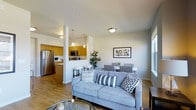 This single-floor residence offers a thoughtfully designed open-concept layout that integrates the living, dining, and kitchen areas into one cohesive and functional space. The living room serves as the central hub, featuring light hardwood-style flooring that flows throughout the unit, neutral-toned walls, and ample natural light streaming in through large windows. A comfortable seating arrangement anchors the space, complemented by a glass-top coffee table and tasteful wall art that adds a modern touch to the ambiance. Adjacent to the living area, the dining space accommodates a full table and chairs, making it ideal for everyday meals or entertaining guests. The kitchen is partially separated by a structural column and a breakfast bar with seating, offering both visual distinction and an open feel. Warm honey-toned wood cabinetry lines the kitchen walls, paired with stainless steel appliances — including a full-size refrigerator, microwave, and range — for a contemporary and practical finish. Pendant lighting above the breakfast bar adds a stylish accent, while recessed and flush-mount ceiling lights ensure the space is well-lit throughout. The overall ambiance is clean, modern, and inviting, with a cohesive design palette that balances functionality with aesthetic appeal.