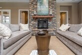 Located at 700 SR-96 E in Bonaire, Georgia, this single-floor space is designed with a refined, welcoming aesthetic that blends warmth and sophistication throughout. The centerpiece of the main lounge area is a striking floor-to-ceiling stacked-stone fireplace, topped with a rich wood mantel and a large framed artwork, creating a commanding focal point. Surrounding seating — including plush sofas and patterned accent chairs — is arranged to encourage relaxed gathering and conversation.

Overhead, an impressive coffered ceiling with dark wood beams and recessed lighting adds architectural depth and a sense of grandeur to the space. Tall, multi-pane glass doors along the exterior wall flood the interior with natural light while offering views of the outdoor surroundings, enhancing the open, airy feel of the room.

Rich hardwood flooring runs throughout, grounding the warm, neutral color palette of the walls and furnishings. A secondary area features a wall-mounted flat-screen television alongside a dark wood display bookcase, offering an additional comfortable retreat. Connecting hallways lead to supplementary rooms, suggesting a well-organized and functional layout. Overall, the space exudes an inviting, upscale ambiance suited for community gathering or hospitality use.