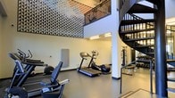 This contemporary two-story fitness center is thoughtfully designed to accommodate a full range of workout activities across two connected levels. The ground floor serves as the primary exercise area, equipped with cardio machines including treadmills and an elliptical, alongside strength training equipment and weight benches. The space is anchored by impressively high, double-height ceilings that create an open, airy atmosphere and contribute to a sense of spaciousness throughout. A standout design feature is the large-scale geometric wall mural that spans a significant portion of the upper wall, composed of a dynamic triangular pattern in a palette of black, white, and muted tones, adding a bold artistic focal point to the functional space. A sleek black spiral staircase connects the ground level to the upper floor mezzanine, which overlooks the main workout area through a railed balcony. The flooring on the main level is a durable rubberized surface well-suited for exercise use. Overhead lighting provides consistent illumination throughout. The overall ambiance balances industrial-modern aesthetics with practical fitness functionality, making it both visually engaging and purpose-driven.