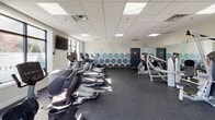 Located at 321 S Broad St in Elizabeth, NJ, this fitness center offers a well-organized and comprehensively equipped exercise facility across a single floor. The space features a clean, modern aesthetic with neutral-toned walls, a drop tile ceiling with recessed panel lighting, and durable rubber flooring throughout — ideal for high-traffic athletic use.

The cardio zone is generously outfitted with treadmills, elliptical trainers, and stationary bikes, including upright and recumbent options, all positioned to take advantage of the natural light streaming through multiple large windows. A wall-mounted flat-screen television provides entertainment for cardio sessions.

The strength and functional training area houses a cable resistance machine system by Hoist, along with a variety of free weights, medicine balls, stability balls, and a BOSU trainer. Full-length mirrors line portions of the walls, enhancing both the sense of space and usability for form-focused training.

A striking honeycomb geometric mural in blue tones adds a vibrant design element to one wall, giving the space a modern, energetic character. The entrance features glass double doors that allow views to the exterior, contributing to the open and welcoming atmosphere. The fitness center is well-lit, thoughtfully arranged, and equipped to support a wide range of workout routines.