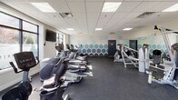 Located at 321 S Broad St in Elizabeth, NJ, this fitness center offers a well-organized and comprehensively equipped exercise facility across a single floor. The space features a clean, modern aesthetic with neutral-toned walls, a drop tile ceiling with recessed panel lighting, and durable rubber flooring throughout — ideal for high-traffic athletic use.

The cardio zone is generously outfitted with treadmills, elliptical trainers, and stationary bikes, including upright and recumbent options, all positioned to take advantage of the natural light streaming through multiple large windows. A wall-mounted flat-screen television provides entertainment for cardio sessions.

The strength and functional training area houses a cable resistance machine system by Hoist, along with a variety of free weights, medicine balls, stability balls, and a BOSU trainer. Full-length mirrors line portions of the walls, enhancing both the sense of space and usability for form-focused training.

A striking honeycomb geometric mural in blue tones adds a vibrant design element to one wall, giving the space a modern, energetic character. The entrance features glass double doors that allow views to the exterior, contributing to the open and welcoming atmosphere. The fitness center is well-lit, thoughtfully arranged, and equipped to support a wide range of workout routines.