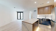 Located at 1923 Broadway Blvd in Kansas City, Missouri, this thoughtfully designed one-bedroom, one-bathroom residence offers a compelling blend of modern aesthetics and functional living. The open-concept kitchen serves as a centerpiece, showcasing two-tone cabinetry with warm wood uppers and bold navy blue lower cabinets, complemented by under-cabinet lighting. A striking quartz-topped island with a marble-effect waterfall base anchors the space, while stainless steel appliances and a marble-style backsplash complete the upscale finish. Light hardwood flooring runs throughout the main living area, enhancing the bright, airy atmosphere created by recessed lighting. The bathroom is a design standout, featuring marble-look tile flooring, a sleek vanity with gold hardware accents, matte black fixtures, and a glass-enclosed shower adorned with deep navy vertical tile from floor to ceiling. The bedroom offers plush carpeting, a ceiling fan, and a large window framing views of the Kansas City skyline and surrounding cityscape, filling the room with natural light. A dedicated closet and hallway complete the layout, providing practical storage within a well-appointed, contemporary urban residence.