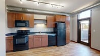 This efficiently designed 2-bedroom, 1-bathroom unit offers a well-organized layout across a single floor. The open kitchen serves as a central feature, equipped with warm medium-tone wood cabinetry, dark countertops, and a full suite of black appliances including a refrigerator, dishwasher, stove, and built-in microwave. Track lighting and exposed industrial-style ductwork along the ceiling add a contemporary urban character to the space. Light hardwood flooring runs throughout the main living and kitchen areas, contributing to a bright and cohesive feel. Large floor-to-ceiling windows and a glass-paneled door flood the living area with natural light and provide direct access to an outdoor area, enhancing the sense of openness. The hallway connects the main living space to the bedrooms and bathroom. Both bedrooms feature plush carpeting and ample closet space with wire shelving systems — the primary bedroom includes a sliding-door closet, while the second bedroom offers its own storage. The bathroom is finished with light tile flooring, a wood vanity with a white countertop, and a full bathtub with a curved shower curtain rod. Neutral wall tones throughout create a clean, functional, and move-in-ready environment.