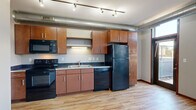 This efficiently designed 2-bedroom, 1-bathroom unit offers a well-organized layout across a single floor. The open kitchen serves as a central feature, equipped with warm medium-tone wood cabinetry, dark countertops, and a full suite of black appliances including a refrigerator, dishwasher, stove, and built-in microwave. Track lighting and exposed industrial-style ductwork along the ceiling add a contemporary urban character to the space. Light hardwood flooring runs throughout the main living and kitchen areas, contributing to a bright and cohesive feel. Large floor-to-ceiling windows and a glass-paneled door flood the living area with natural light and provide direct access to an outdoor area, enhancing the sense of openness. The hallway connects the main living space to the bedrooms and bathroom. Both bedrooms feature plush carpeting and ample closet space with wire shelving systems — the primary bedroom includes a sliding-door closet, while the second bedroom offers its own storage. The bathroom is finished with light tile flooring, a wood vanity with a white countertop, and a full bathtub with a curved shower curtain rod. Neutral wall tones throughout create a clean, functional, and move-in-ready environment.