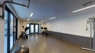 This fitness facility offers a clean, contemporary workout environment designed to support a variety of exercise routines. The space features a warm wood-tone floor that provides both durability and comfort underfoot, complemented by two-tone walls — crisp white on the upper portion and a cool charcoal gray on the lower half — creating a sleek, modern aesthetic throughout.

Bright linear LED lighting fixtures are mounted flush along the ceiling, bathing the entire space in even, high-quality illumination ideal for an active environment. The ceiling height contributes to an open, airy feel, making the room feel spacious and unrestricted.

The main exercise area is outfitted with stationary cycling bikes equipped with digital display screens, suitable for guided or self-directed cardio workouts. A stability ball and additional equipment are also present, supporting a range of fitness activities. The open floor plan allows for flexible use of the space, accommodating group fitness classes or individual training sessions with ease.

A set of frosted glass double doors serves as the main entrance, offering both privacy and a refined architectural touch. An additional interior door provides further access or utility. Safety signage, including an illuminated exit sign, ensures the space meets practical standards. Overall, this fitness facility is thoughtfully designed to deliver a functional, motivating, and comfortable workout experience.
