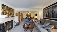 This single-floor residence offers a warm and inviting layout designed for comfortable everyday living. The living room serves as the heart of the home, featuring a tile-surround fireplace that anchors the space with a cozy focal point. Plush carpeting in neutral tones extends throughout, complementing the soft, earthy color palette that runs across the interior. Two generously sized sofas in muted gray provide ample seating, arranged around a classic round wood coffee table with decorative ironwork detailing. A ceiling fan overhead offers both function and a subtle design element, while tasteful artwork and decorative accents — including framed prints and sculptural pieces — add personality to the walls and surfaces. The living area opens seamlessly into a dining space beyond, creating an easy, connected flow ideal for entertaining or daily living. Interior doors visible from the main living space suggest well-organized access to additional rooms. Natural light filters through the space, enhancing the warm, welcoming ambiance. Overall, the home presents a thoughtfully arranged, move-in-ready interior with a classic aesthetic and practical, comfortable layout throughout its single level.