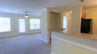 This single-floor residence features an open-concept layout that connects the kitchen and living areas seamlessly. The kitchen includes a raised breakfast bar with granite-style countertops, offering both a functional workspace and a casual dining option. A built-in nook adjacent to the bar provides additional storage or display space. The living area is finished with plush wall-to-wall carpeting and is bathed in natural light from a large set of windows along the exterior wall. A ceiling fan with an integrated light fixture is centrally positioned, contributing to both comfort and ambient lighting. Neutral yellow-toned walls and white crown molding trim throughout give the space a clean, cohesive aesthetic. A doorway visible toward the rear of the unit suggests access to additional rooms beyond the main living area. The overall ambiance is bright, airy, and well-maintained, with a practical floor plan suited for everyday living.