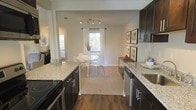 This single-floor residence offers a thoughtfully designed open-concept layout that seamlessly connects the kitchen and dining areas, creating a cohesive and inviting living environment. The kitchen is a standout feature, showcasing rich dark cabinetry paired with light granite countertops that provide a striking contrast. A smooth-surface electric range and stainless steel sink complement the space, while track lighting overhead ensures ample illumination for both cooking and entertaining. Rich hardwood flooring runs through the kitchen, adding warmth and a sense of continuity. The dining area transitions naturally from the kitchen, featuring neutral carpeting that softens the space. A bright window dressed with white curtains draws in natural light, creating an airy atmosphere around the dining set. The walls are finished in a soft, neutral tone that enhances the brightness of the space and complements the dark cabinetry. Framed artwork adorns the walls, adding a curated, polished touch. Additional windows visible throughout suggest generous natural light across the entire floor plan. The overall design balances modern aesthetics with functional living, making this a well-appointed and comfortable space ideal for everyday living and entertaining alike.