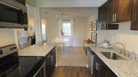 This single-floor residence offers a thoughtfully designed open-concept layout that seamlessly connects the kitchen and dining areas, creating a cohesive and inviting living environment. The kitchen is a standout feature, showcasing rich dark cabinetry paired with light granite countertops that provide a striking contrast. A smooth-surface electric range and stainless steel sink complement the space, while track lighting overhead ensures ample illumination for both cooking and entertaining. Rich hardwood flooring runs through the kitchen, adding warmth and a sense of continuity. The dining area transitions naturally from the kitchen, featuring neutral carpeting that softens the space. A bright window dressed with white curtains draws in natural light, creating an airy atmosphere around the dining set. The walls are finished in a soft, neutral tone that enhances the brightness of the space and complements the dark cabinetry. Framed artwork adorns the walls, adding a curated, polished touch. Additional windows visible throughout suggest generous natural light across the entire floor plan. The overall design balances modern aesthetics with functional living, making this a well-appointed and comfortable space ideal for everyday living and entertaining alike.