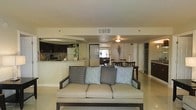 This single-floor residence features an open-concept layout that integrates the living, dining, and kitchen areas into one cohesive space. The living area is anchored by a light-toned sofa accented with patterned throw pillows in muted blues and grays, providing a comfortable and stylish focal point. The neutral-toned flooring and cream-colored walls contribute to a bright, airy ambiance throughout.

The kitchen is positioned to the rear left, featuring dark cabinetry with a contrasting light granite or stone countertop, a white refrigerator, and a tiled backsplash. A breakfast bar or peninsula separates the kitchen from the rest of the open space, making it ideal for casual dining or entertaining. Recessed and flush-mounted ceiling fixtures provide consistent lighting throughout.

The dining area is situated centrally, furnished with a dark wood table and matching chairs, bridging the kitchen and living spaces naturally. A raised ceiling detail with a circular tray design adds architectural interest overhead. Overall, the space presents a clean, well-organized layout suited for comfortable everyday living.