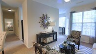 This single-story space showcases a well-appointed living area designed with a contemporary aesthetic. The room features neutral gray walls that serve as a versatile backdrop, complemented by light-toned carpeting that adds warmth underfoot. Vaulted or elevated ceilings contribute a sense of openness and airiness to the interior.

A standout decorative element is the starburst mirror mounted prominently on the accent wall, adding a bold focal point to the space. Beneath it, a dark-finished console table with drawer storage and open shelving provides both style and function. The furnishings include patterned accent chairs with intricate designs, layered over a geometric area rug, creating visual interest throughout the room.

Natural light flows generously through large windows dressed with blinds and sheer curtains, illuminating the space and offering a bright, welcoming atmosphere. The layout flows seamlessly, with a secondary seating or dining area visible toward the rear, also benefiting from ample window light. The overall ambiance is modern and curated, balancing decorative flair with comfortable, functional living.