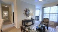 This single-story space showcases a well-appointed living area designed with a contemporary aesthetic. The room features neutral gray walls that serve as a versatile backdrop, complemented by light-toned carpeting that adds warmth underfoot. Vaulted or elevated ceilings contribute a sense of openness and airiness to the interior.

A standout decorative element is the starburst mirror mounted prominently on the accent wall, adding a bold focal point to the space. Beneath it, a dark-finished console table with drawer storage and open shelving provides both style and function. The furnishings include patterned accent chairs with intricate designs, layered over a geometric area rug, creating visual interest throughout the room.

Natural light flows generously through large windows dressed with blinds and sheer curtains, illuminating the space and offering a bright, welcoming atmosphere. The layout flows seamlessly, with a secondary seating or dining area visible toward the rear, also benefiting from ample window light. The overall ambiance is modern and curated, balancing decorative flair with comfortable, functional living.