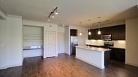 This single-story space in North Richland Hills, Texas, showcases a thoughtfully designed open-concept layout that seamlessly connects the kitchen and living areas. The kitchen is a standout feature, outfitted with dark espresso cabinetry that contrasts elegantly against a white subway tile backsplash and light-toned countertops. A central kitchen island with a built-in sink provides additional workspace and serves as a natural gathering point. Stainless steel appliances, including a refrigerator, stove, and microwave, add a polished, contemporary touch. Pendant lighting hangs above the island, complementing the track lighting installed throughout the main living space to ensure well-distributed illumination. Rich, dark hardwood-style flooring runs continuously across the open area, lending warmth and visual cohesion to the space. A built-in shelving nook near a paneled interior door offers practical display or storage options, while the neutral wall tones keep the overall atmosphere bright and inviting. The open floor plan enhances the sense of spaciousness, making this a functional and modern living environment.