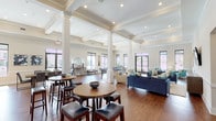 This well-appointed single-floor clubhouse offers a thoughtfully designed layout that seamlessly connects multiple social and functional zones. The main lounge area commands attention with its soaring coffered ceilings, large structural columns, and abundant natural light streaming through an array of windows. Rich hardwood flooring flows throughout, providing a warm and cohesive aesthetic across the entire space. The lounge is furnished with plush navy blue sofas and teal accent chairs, complemented by a wall-mounted flat-screen television and a stylish media console, creating a comfortable and inviting gathering area. Tall bar-height tables and stools offer additional casual seating near the windows. A dedicated dining area accommodates group gatherings at a substantial rectangular table surrounded by quilted upholstered chairs, with views toward an outdoor pool area through large glass doors. The fully equipped kitchen features dark textured cabinetry, crisp white quartz countertops, a subway tile backsplash, stainless steel appliances including a gas range and dishwasher, and a breakfast bar with seating. A billiards table rounds out the amenities, adding a recreational dimension to this versatile community space.