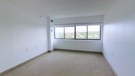 This single-floor space offers a clean, minimalist interior defined by its most striking feature: a large, multi-pane window spanning a considerable portion of one wall. Positioned at an elevated level, the window frames panoramic views of the surrounding urban landscape, including nearby buildings and a canopy of mature trees stretching into the distance, filling the room with abundant natural light throughout the day.

The interior is finished with neutral, light-toned carpeting that extends across the open floor area, complementing the crisp white walls and contributing to an overall sense of brightness and openness. The ceiling is smooth and white, with visible structural beams adding a subtle architectural detail overhead. A ceiling-mounted ventilation duct is integrated discreetly near the window, supporting climate control throughout the space.

The walls are largely unadorned, providing a versatile, blank-canvas layout suitable for a variety of uses. Electrical outlets and light switch plates are positioned along the walls. The baseboard trim is clean and minimal, consistent with the space's understated aesthetic. Overall, the space presents a functional, light-filled environment with a notable connection to the outdoor cityscape.