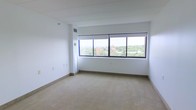 This single-floor space offers a clean, minimalist interior defined by its most striking feature: a large, multi-pane window spanning a considerable portion of one wall. Positioned at an elevated level, the window frames panoramic views of the surrounding urban landscape, including nearby buildings and a canopy of mature trees stretching into the distance, filling the room with abundant natural light throughout the day.

The interior is finished with neutral, light-toned carpeting that extends across the open floor area, complementing the crisp white walls and contributing to an overall sense of brightness and openness. The ceiling is smooth and white, with visible structural beams adding a subtle architectural detail overhead. A ceiling-mounted ventilation duct is integrated discreetly near the window, supporting climate control throughout the space.

The walls are largely unadorned, providing a versatile, blank-canvas layout suitable for a variety of uses. Electrical outlets and light switch plates are positioned along the walls. The baseboard trim is clean and minimal, consistent with the space's understated aesthetic. Overall, the space presents a functional, light-filled environment with a notable connection to the outdoor cityscape.