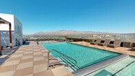 Perched atop a high-rise building at 916 E Speedway Blvd in Tucson, Arizona, this rooftop amenity deck offers an exceptional outdoor living experience with breathtaking, unobstructed views of the surrounding Sonoran Desert cityscape and the rugged mountain ranges beyond. The centerpiece of the space is a generously sized resort-style swimming pool with crystal-clear turquoise water, accessible via wide entry steps with stainless steel handrails. The pool deck is finished in warm-toned large-format tiles that complement the desert environment. Surrounding the pool is an expansive sun deck furnished with rows of sleek, cushioned chaise loungers positioned to maximize both sunbathing and scenic views. Shaded retreats are provided by several structured wooden pergola cabanas, offering comfortable seating for relaxation away from direct sun. A separate upper terrace area features a patch of lush green lawn alongside additional lounge seating and outdoor dining, providing a versatile space for social gatherings. Glass perimeter railings ensure unobstructed sightlines across the Tucson skyline and toward the Santa Catalina Mountains, making this rooftop deck a standout destination for both leisure and entertaining under Arizona's abundant sunshine.